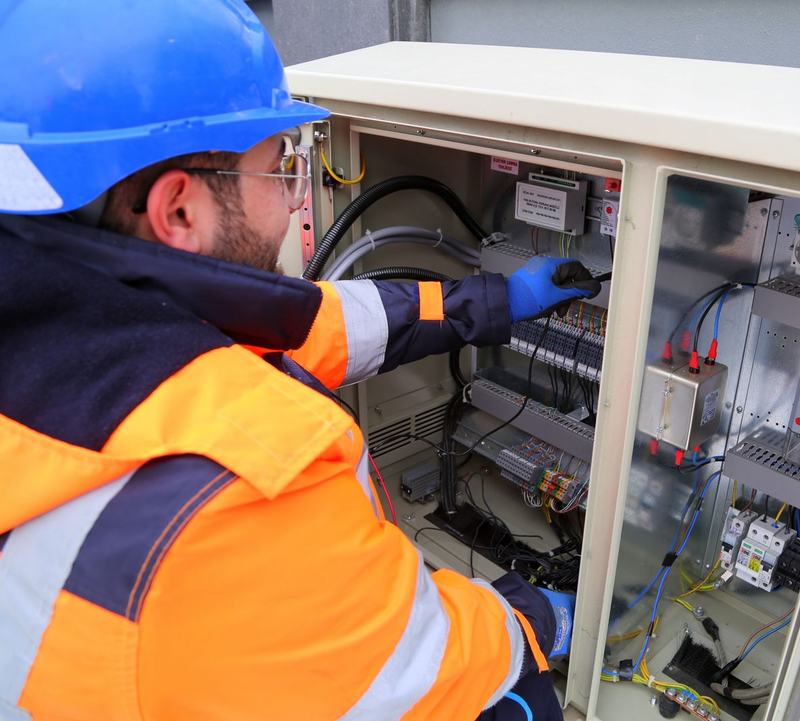 Electrician in hard hat, hi-vis and gloves testing terminations in an outdoor control cabinet