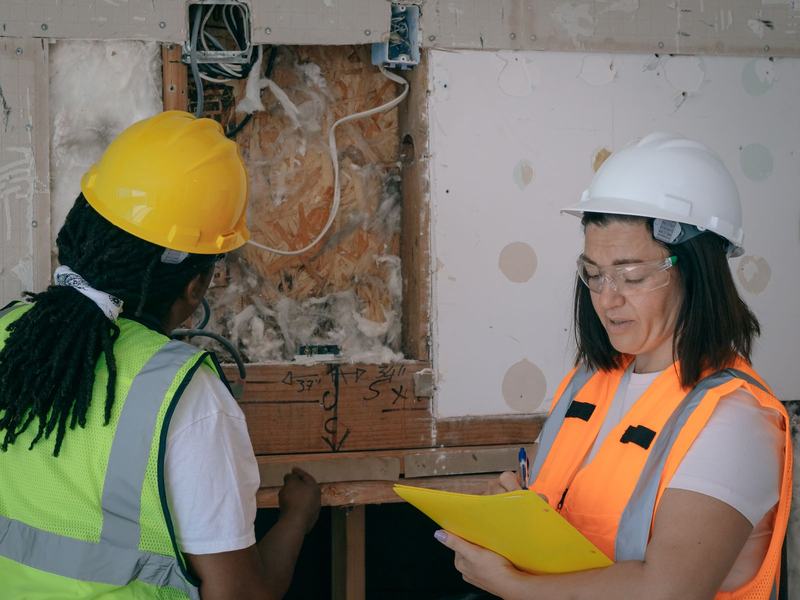 Two engineers in hard hats and hi-vis recording an electrical first-fix inspection on a clipboard
