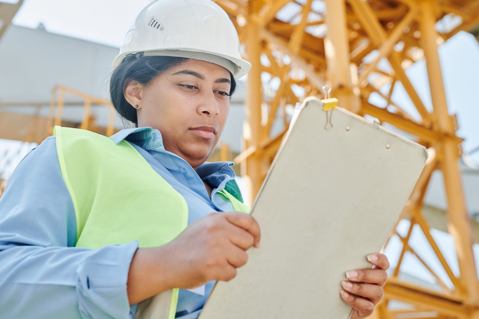Engineer in hard hat and hi-vis completing an inspection record on a clipboard at the base of a tower crane